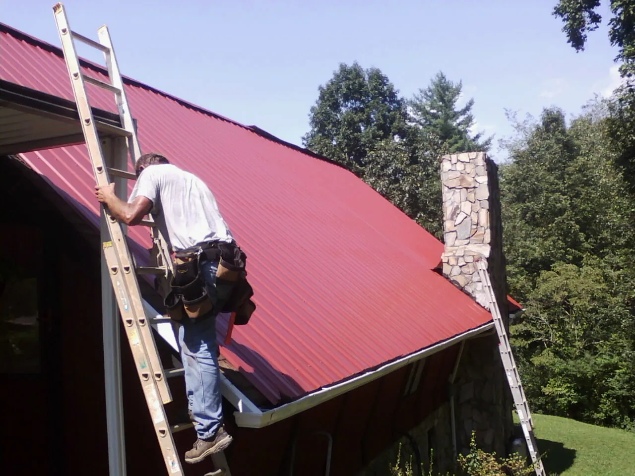 Roofer on a ladder beside a red metal roof and stone chimney — Soffit & Fascia Repair by Roofing Brothers in Port Washington